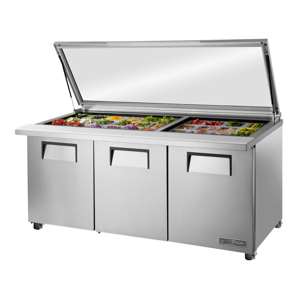 A True 3 door refrigerated sandwich prep table with a hinged glass lid on a counter with stainless steel trays inside.