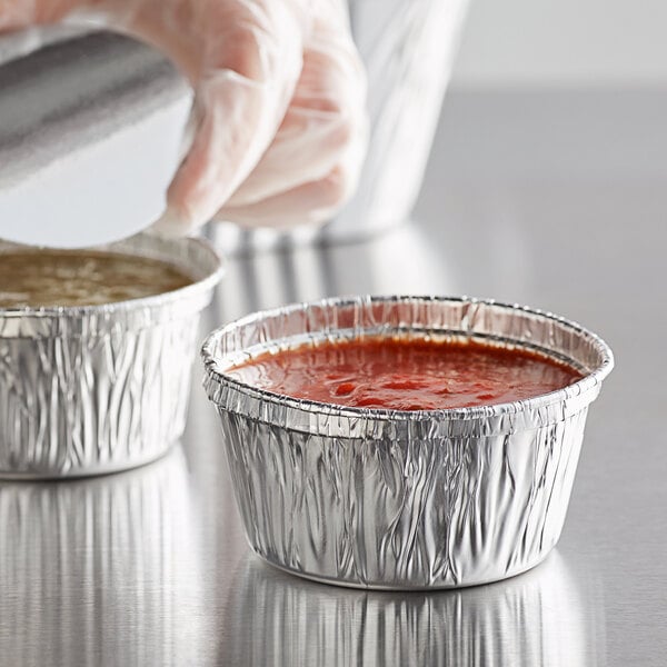 A gloved hand pouring red sauce into a 4 oz. foil ramekin.