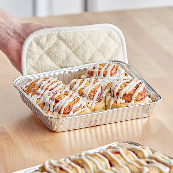 A hand holding a Baker's Lane foil tray of cinnamon rolls with white frosting.