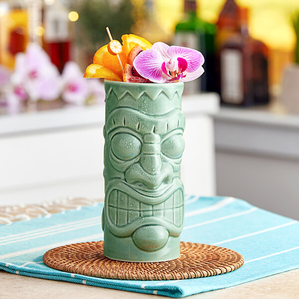 A green ceramic Acopa tiki mug with orange flowers on a woven mat.