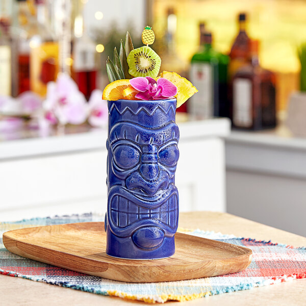 A blue Acopa ceramic tiki mug with fruit and flowers on a wooden tray.