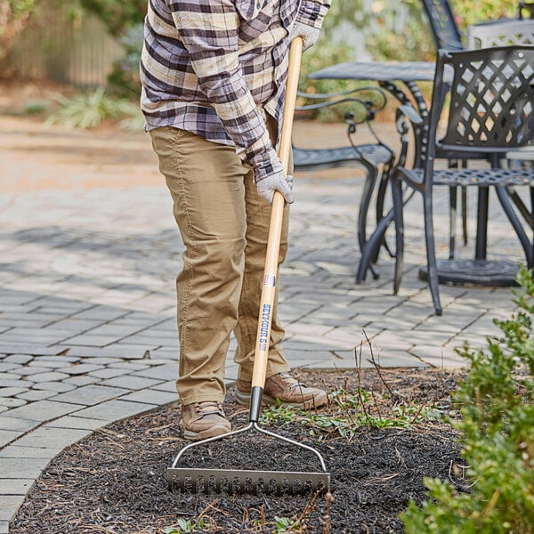 A person using a Seymour steel bow rake with a wooden handle to clean the ground.