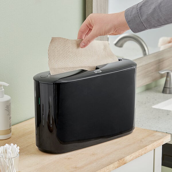 A person using a Tork black countertop multifold hand towel dispenser.