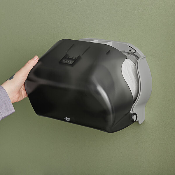 A hand holding a black plastic Tork double roll toilet paper dispenser.