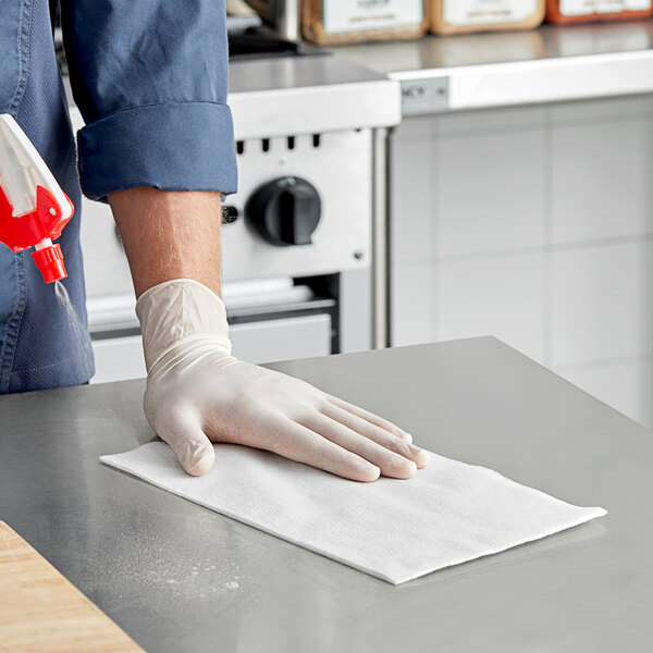 A person wearing a glove and using a Tork white food service towel to clean a surface.