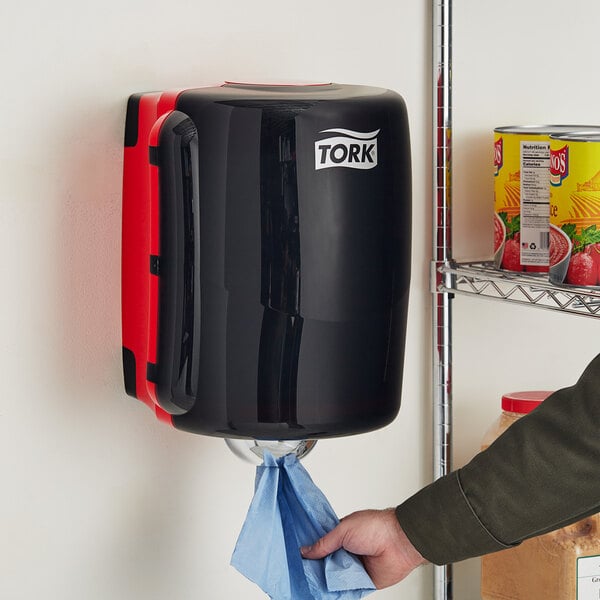 A person using a red and black Tork Maxi center pull towel dispenser.