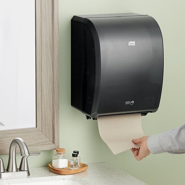 A hand pulling a paper towel out of a black Tork electronic hand towel dispenser.
