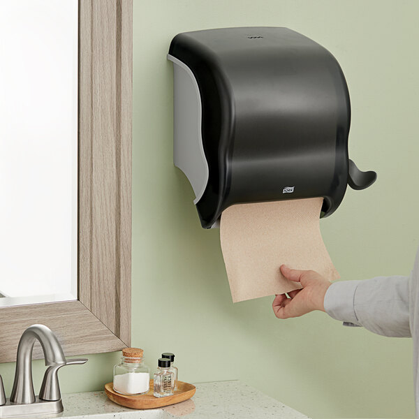 A hand holding a roll of paper towels in a Tork Black Lever Activated Roll Hand Towel Dispenser.