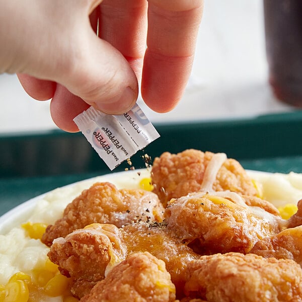 A finger holding a Black Pepper portion packet over a plate of fried chicken.