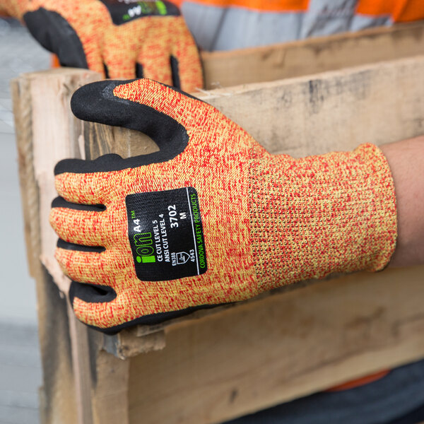 A person wearing Cordova orange cut-resistant gloves with black sandy nitrile palms holding a piece of wood.