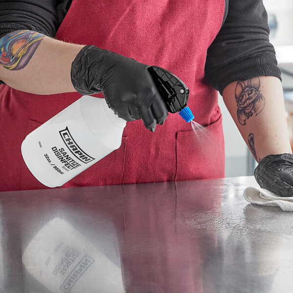 A person wearing gloves and a black apron spraying a metal surface with a white Chapin spray bottle.