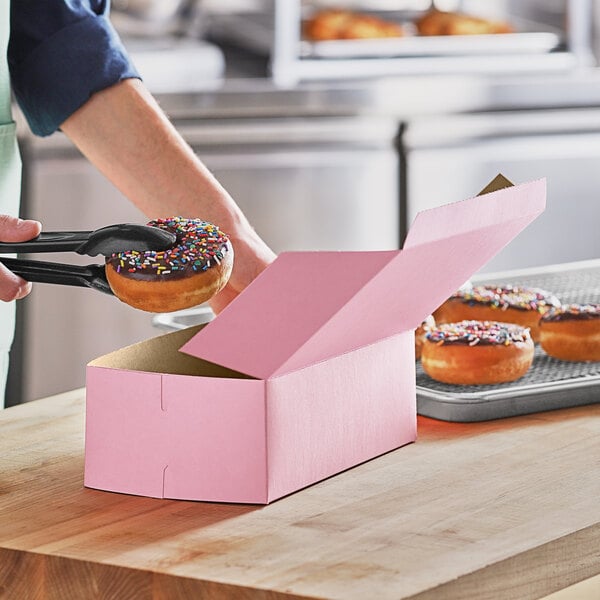 A person holding a Baker's Mark pink donut box with a donut with sprinkles inside.