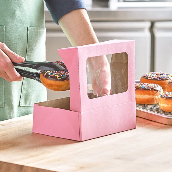A hand using tongs to pick up a donut with pink sprinkles in a bakery display.