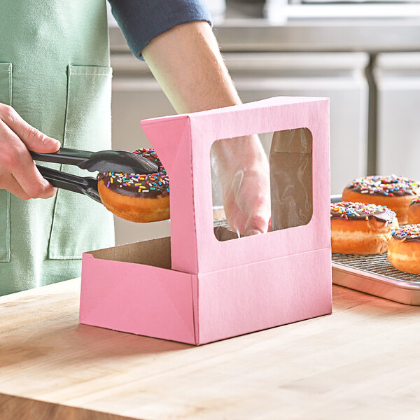A hand using tongs to pick up a donut with pink sprinkles from a Baker's Mark bakery box with a window.