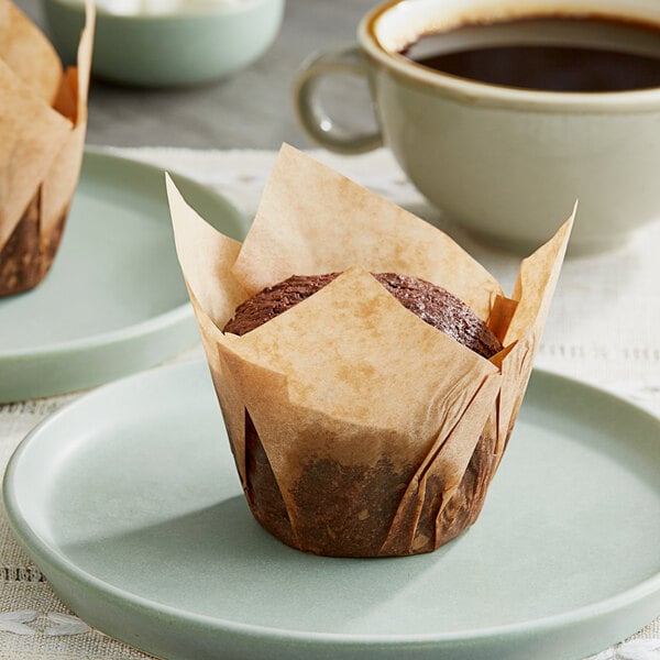 Two Baker's Mark unbleached natural kraft tulip baking cups holding chocolate muffins on a plate next to a cup of coffee.