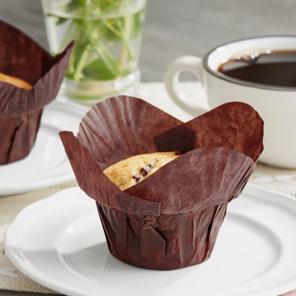 Two Baker's Mark chocolate brown muffins in large rounded wrappers on a plate.