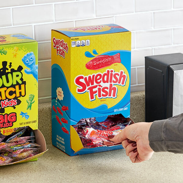 A hand reaching out to a yellow Swedish Fish candy box on a white counter.