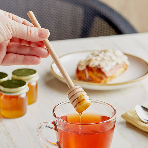 A hand holding a Fox Run wooden honey dipper over a glass of honey.