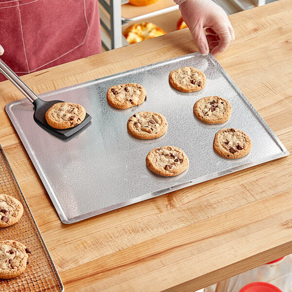 A hand in a glove using a spatula to put a chocolate chip cookie on a DoughMakers pebbled baking sheet.