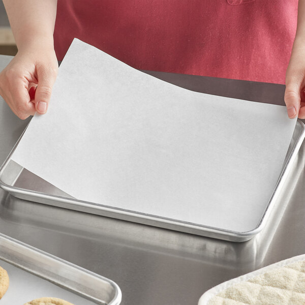 A person holding a Baker's Lane Quilon-coated parchment paper sheet over a tray of cookies.