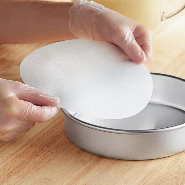 A person's gloved hands placing a round white Baker's Lane pan liner in a round metal pan.