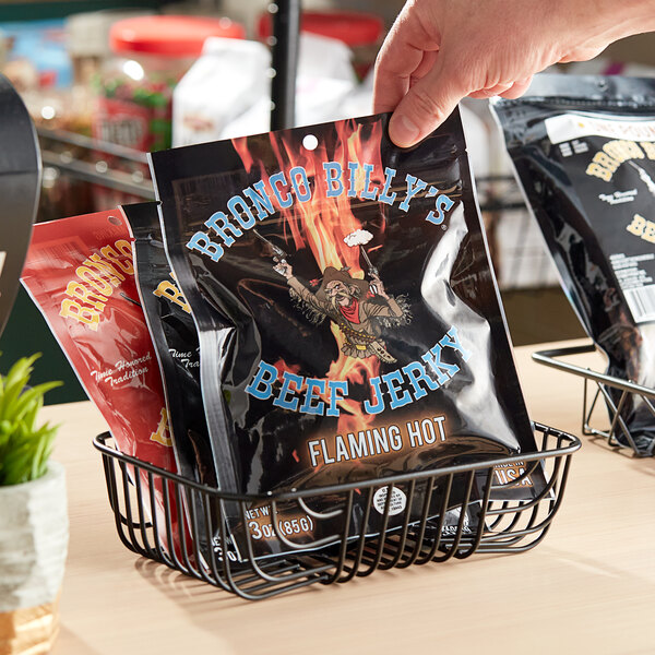 A hand holding a black bag of Bronco Billy's Flaming Hot Beef Jerky with a cartoon character on the label.
