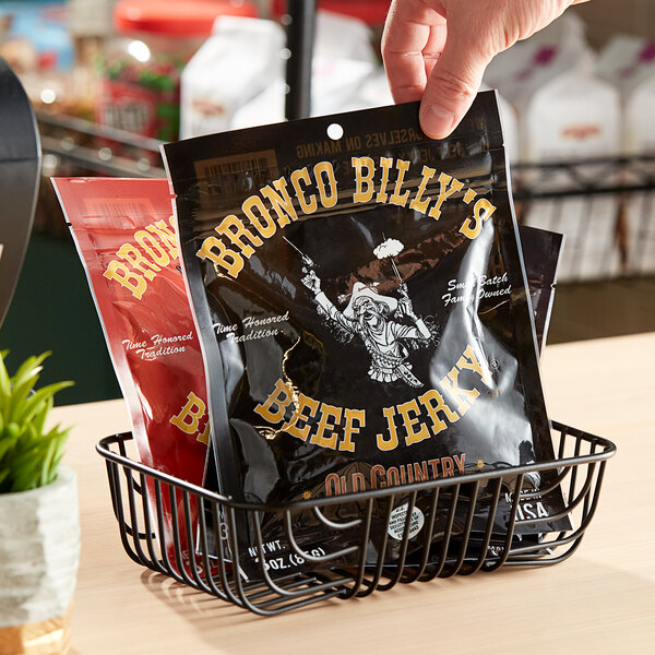 A hand holding a bag of Bronco Billy's Old Country Beef Jerky in a black basket.
