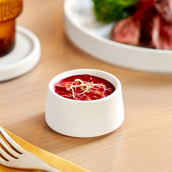 A small, bright white porcelain sauce cup filled with red sauce and garnished with sprouts.