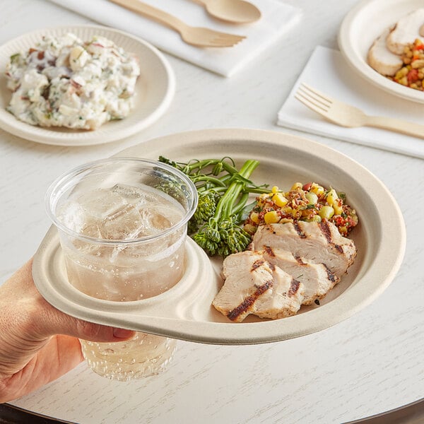 A hand holding a World Centric compostable plate of food over a glass of liquid.