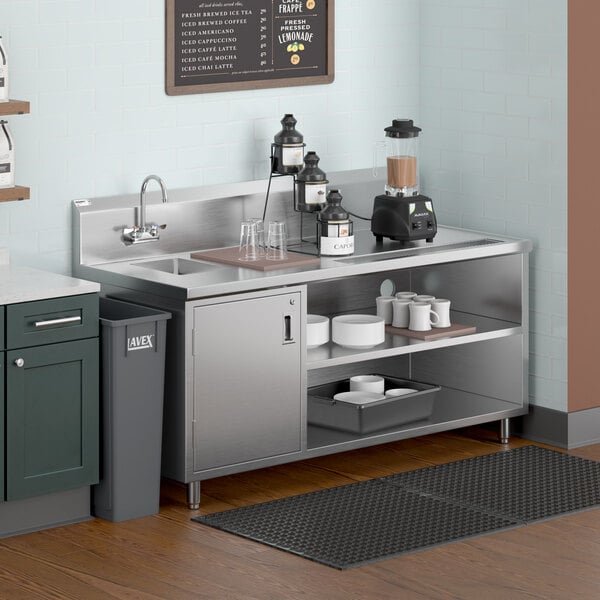 A stainless steel beverage station with a left-side sink, gooseneck faucet, open shelves holding cups and dishes, a blender, and condiment dispensers, set against a light blue tiled wall.