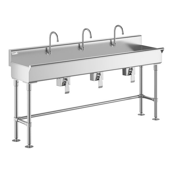 A large stainless steel sink with three gooseneck faucets and knee-operated controls, supported by metal legs.