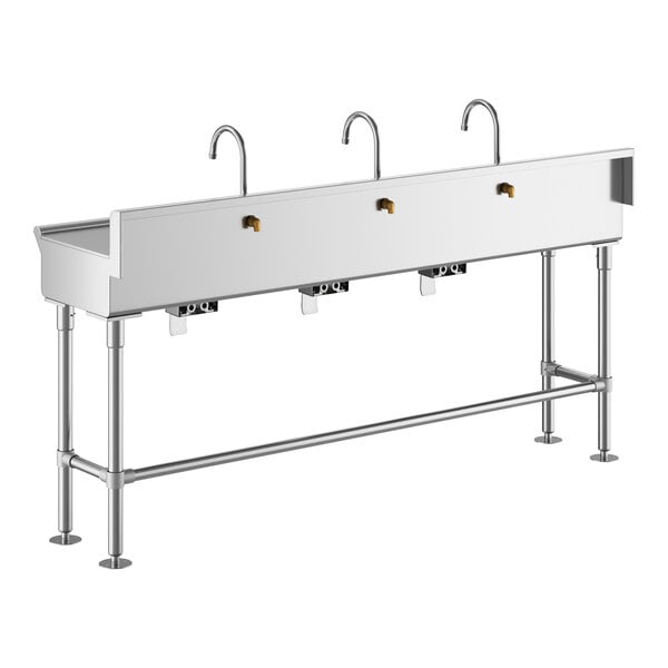 A long, stainless steel hand sink with three gooseneck faucets and knee-operated controls, supported by metal legs.