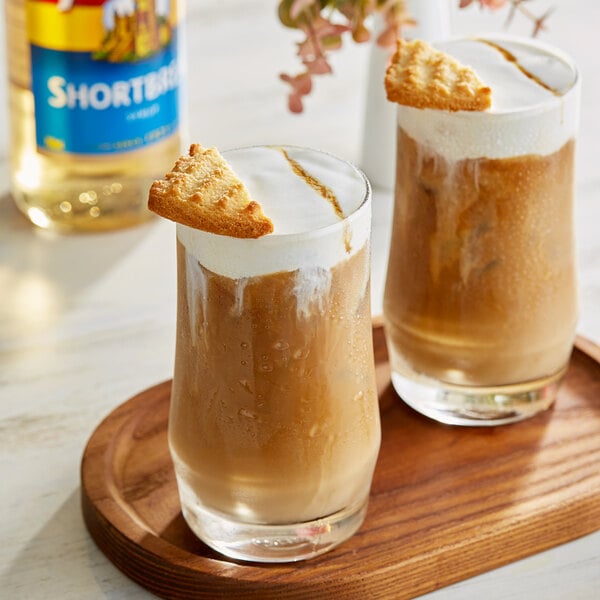 A bottle of Torani Shortbread Flavoring Syrup next to two glasses of brown liquid with a cookie on top.