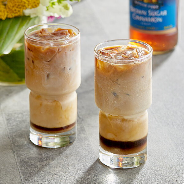 Two glasses of iced coffee with a bottle of Torani Sugar-Free Brown Sugar Cinnamon Flavoring Syrup.