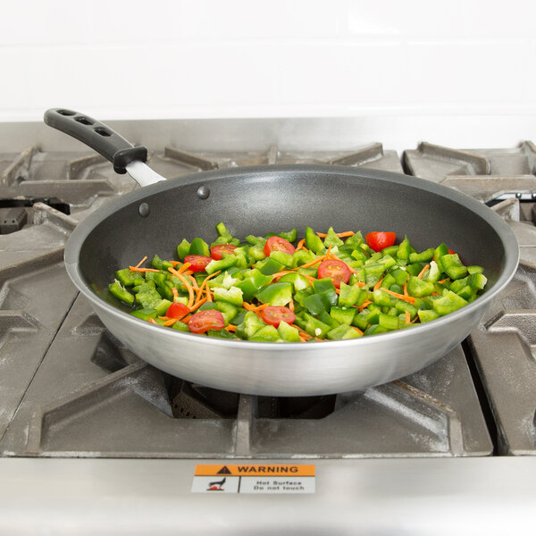 A Vollrath Wear-Ever non-stick fry pan with a black handle full of vegetables on a stove.
