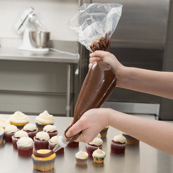 A person uses an Ateco clear disposable pastry bag to pipe brown chocolate frosting on a cupcake.