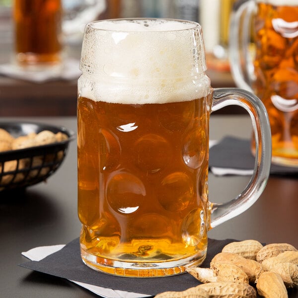 A Libbey glass mug of beer with peanuts on a table.