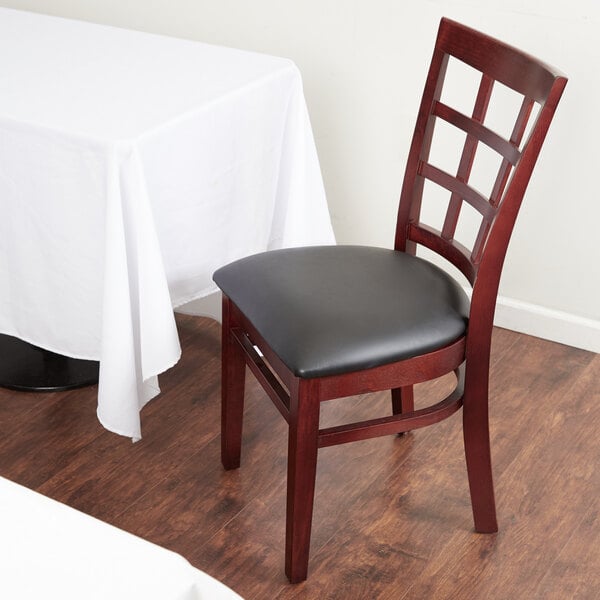 A Lancaster Table & Seating mahogany wood chair with a black vinyl cushion next to a white table.