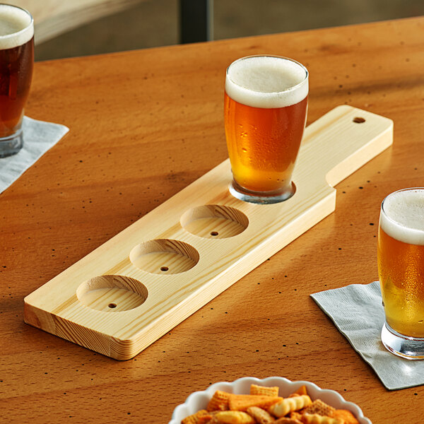 An Acopa natural wood flight paddle with glasses of beer and snacks on a table.