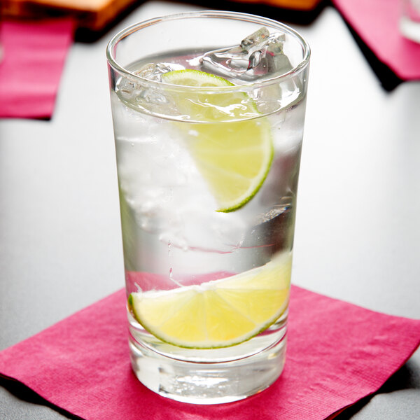 A Libbey customizable highball glass filled with water, ice, and lime slices on a pink napkin.