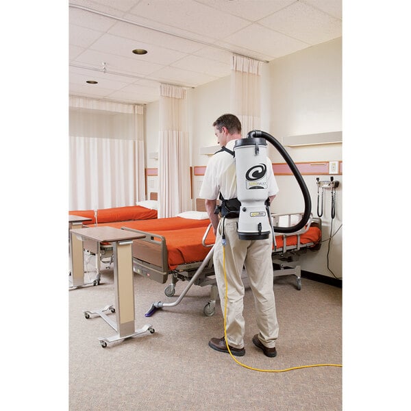 A man in a white suit using a ProTeam LineVacer backpack vacuum to clean a hospital room.