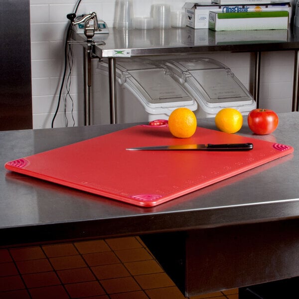 A San Jamar red cutting board with a knife and fruits on a counter.