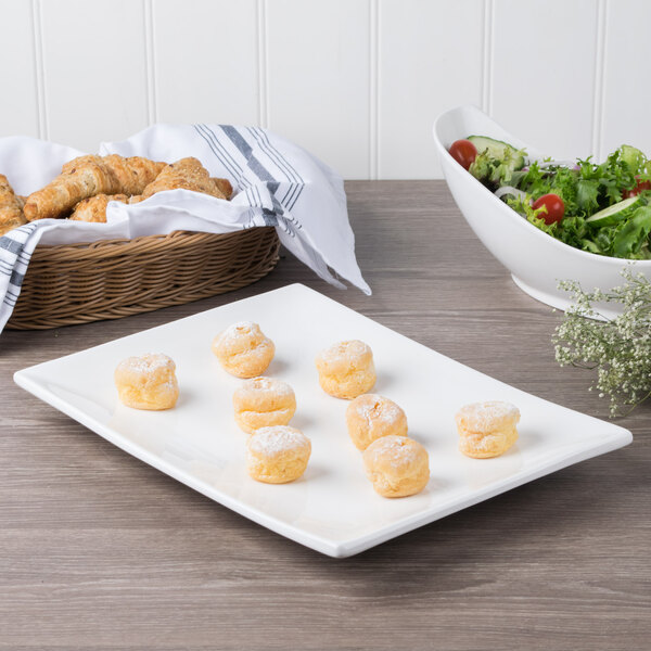 A rectangular white porcelain plate with food on a table next to a bowl of croissants.