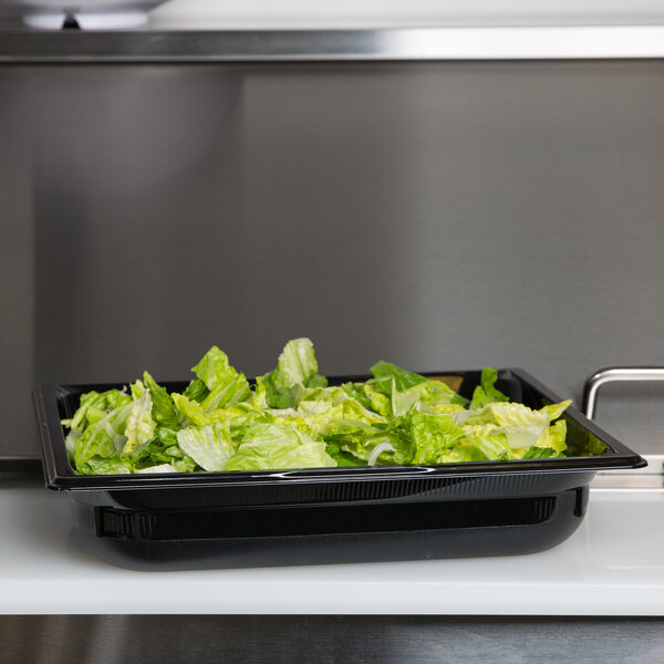 A Vollrath black polycarbonate food pan with lettuce in it on a kitchen counter.
