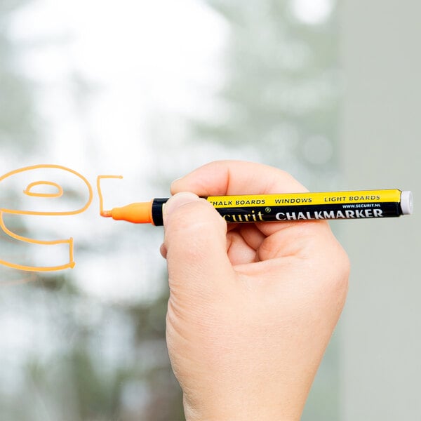 A hand using a yellow American Metalcraft chalk marker to write 'changer' on a window.