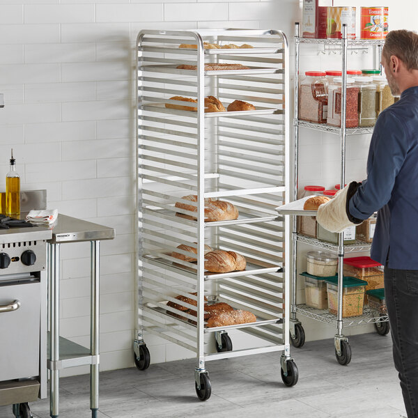 A tall, metal rack with multiple shelves holds several loaves of bread, and a person is placing a tray of bread onto it in a commercial kitchen setting.