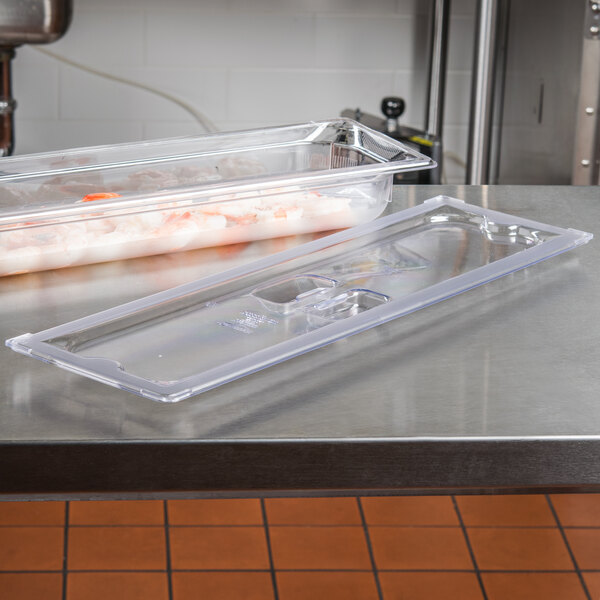 A Vollrath 1/2 size long clear plastic tray with a lid on a table full of food.