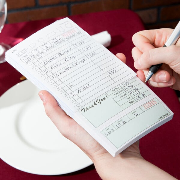 A person holding a green and white carbonless guest check and writing on it.