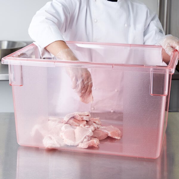 A person in a white coat and gloves putting meat in a Carlisle red food storage container.