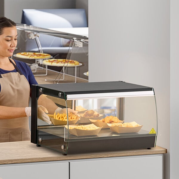 A woman in an apron and gloves putting food in an Avantco countertop food display case.